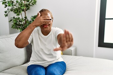 Poster - Mature hispanic woman sitting on the sofa at home covering eyes with hands and doing stop gesture with sad and fear expression. embarrassed and negative concept.