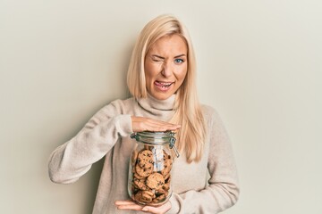 Poster - Young blonde woman holding jar with chocolate chips cookies sticking tongue out happy with funny expression.