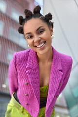Fashionable smiling young woman dressed in pink jacket expresses positive emotions feels good walks in urban place poses outdoors against blurred background. People style and leisure concept