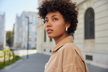 Wall Mural - Sideways shot of thoughtful beautiful young woman with curly bushy hair looks into distance has charming look dressed in beige jacket strolls outdoors against blurred background blank copy space