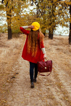 A Girl In A Red Coat And A Yellow Beret With A Suitcase In An Autumn Park. Back To The Camera. 