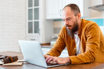 Man use laptop for online work education or shopping. Calm young man working from home. Portrait of bold Bearded Caucasian man Freelancer using his office in kitchen Workspace wooden table at house. 