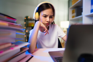 Wall Mural - Stressful  young Asian woman wearing headset and sitting on the chair with a pile of papers document on the table and looking on the computer screen on night.