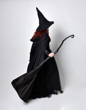 Full Length Portrait Of Dark Haired Woman Wearing  Black Victorian Witch Costume With  Cloak And Pointy Hat.  Standing Pose, Back View,  With  Gestural Hand Movements,  Against Studio Background.