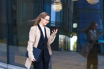 Business woman of Caucasian ethnicity in a coat and a suit with glasses writes SMS messages on the phone near the office building. Business people distance online communication concept