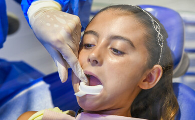 Canvas Print - Young caucasian female with dentist in white latex gloves check condition of her teeth. baby girl in blue dental chair.