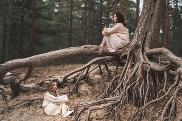 Conceptual photograph of brunettes among forest roots. Beautiful girls sit at the roots of trees and dream. Two friends walk in the woods and talk.