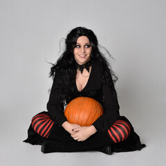 Full length portrait of dark haired woman wearing  black victorian witch costume  sitting pose wit ha pumpkin, with  gestural hand movements,  against studio background.