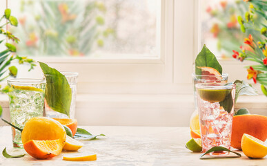 Iced citrus drinks with mint, orange and grapefruit on kitchen table at window with natural light. Homemade healthy lemonade with fruits in summer. Drink background with copy space. Front view.