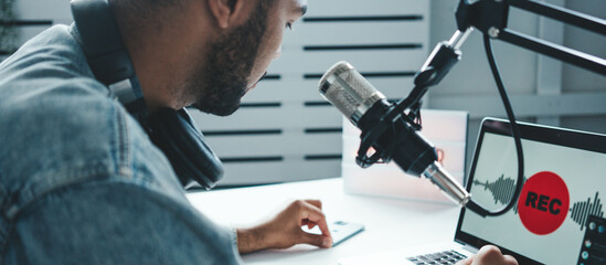 Wall Mural - Handsome mixed race host streaming his audio podcast using microphone and laptop at his homemade broadcast studio