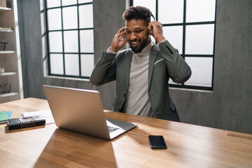 Happy young businessman with smartphone and headset working on laptop indoors in office