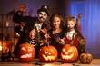 © JenkoAtaman - Young caucasian family mother father and children in Halloween costumes and makeup making scary gesture, saying trick or treat while celebrating together all hallows eve in dark room at home