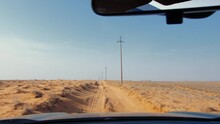 Car Drives Along Road Passing In Desert Area Surrounded By Dry Plants And Electric Power Transmission Poles. View From Auto. Travel By Automobile Through Barren Zone With Sun Scorched Sandy Soil