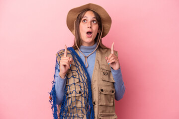 Wall Mural - Young caucasian fisherwoman holding a net isolated on pink background pointing upside with opened mouth.