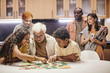 © pressmaster - Grandchildren playing board game together with their grandfather at the table at home with parents standing in the background