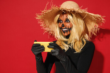 Wall Mural - Young woman with Halloween makeup mask wears straw hat black scarecrow costume using play racing app on mobile cell phone gadget smartphone isolated on plain red background studio Celebration concept