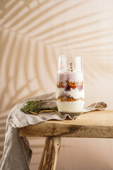 Wall Mural - Tall glass with a layered creamy yoghurt dessert trifle with cookies, amaretti, cherries and thyme on a linen cloth on a rustic wooden table at a tropical setting