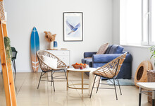 Interior Of Modern Stylish Room With Table, Wicker Armchairs, Sofa And Surfboard