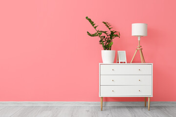 White lamp with frame and houseplant on chest of drawers near pink wall