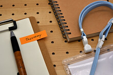 There Is A Sticky Note With The Letters Taxonomy Stamped On The Edge Of A Clipboard Placed Along With A Notebook And Pen.