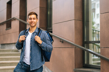 Wall Mural - Medium shot portrait of cheerful handsome young male of food delivery service with large thermo backpack standing posing in office building stairs, selective focus, looking at camera.