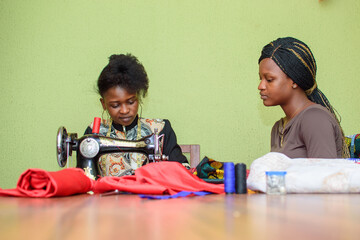 A female African fashion designer or tailor teaching her apprentice how to make clothe using sewing machine in a tailoring workshop