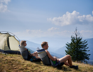 Wall Mural - Two hikers resting in trekking chairs on hill during summer hike in mountains. Pleasant outdoor recreation in mountain spaces. Blue sky with clouds and silhouettes of hills on the background.