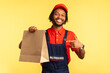 © khosrork - Portrait of happy optimistic workman in uniform and red cap pointing at ordered parcel, delivering food in paper package, post mail services. Indoor studio shot isolated on yellow background.