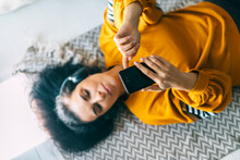 Woman In Casual Clothing With Headphones Lying Down The Floor In Her Living Room And Listening The Music From Her Phone. Leisure Time Of Single Woman In Isolation Streaming Songs From Internet.