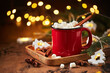Red cup of hot cocoa with marshmallows on wooden table with christmas tree and glowing garland for christmas