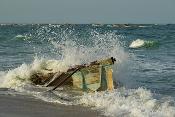 Wall Mural - West Africa. Mauritania. Sea water splashes from the impact of coastal waves on the side of the remains of a broken fishing boat washed up on the Atlantic Ocean near the city of Nouakchott.