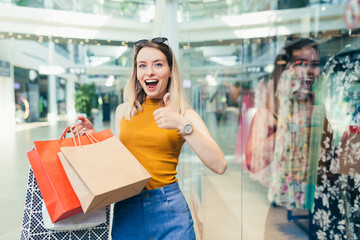 Wall Mural - cheerful young shopaholic woman holding paper bags with purchases and smiling looking at camera dancing on shopping center. Happy lady having fun on black friday joyfully jumps for joy in the mall