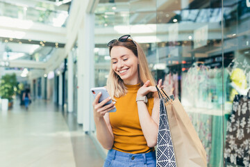 Wall Mural - Young woman consumer in the mall browses chat and uses using a smartphone. female standing with a mobile phone in her hands in shopping center. indoor. happy shopper girl with gift bags make purchases