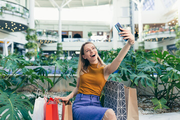 Wall Mural - Young woman consumer in the mall browses chat and uses using a smartphone. female standing with a mobile phone in her hands in shopping center. indoor. happy shopper girl with gift bags make purchases