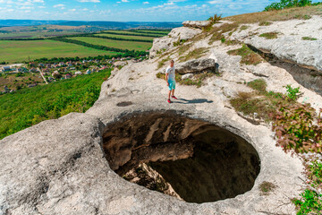 A young man on the edge of a precipice in the form of a huge hole in the ground among green fields and mountains on a sunny summer day. An abnormal cave, a sinkhole in the ground.