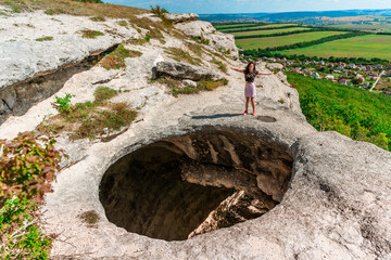 A young woman on the edge of a precipice in the form of a huge hole in the ground among green fields and mountains on a sunny summer day. An abnormal cave, a sinkhole in the ground.