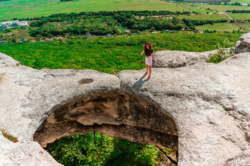 A young woman on the edge of a precipice in the form of a huge hole in the ground among green fields and mountains on a sunny summer day. An abnormal cave, a sinkhole in the ground.