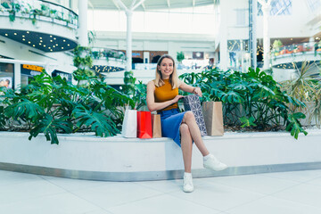 Wall Mural - young happy after shopping woman talking on cell in mall. Satisfied joyful female shopaholic with gift colored paper bags in hands. Mobile phone conversation. Smiling joy Stand ang sincerely tells