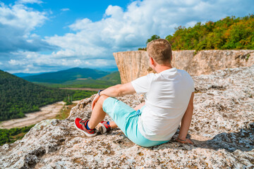 Wall Mural - A young man is sitting on the top of a mountain on a sunny summer day and admiring the mountain landscape. Tourism concept