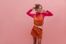 Shot Of Pretty Young Polish Woman With Lovely Smile Dancing Against Pink Background. In Velvet Orange Jumpsuit On Raspberry Sweater, Her Hands Gently Touch Face. Beauty, People Emotion Concept.