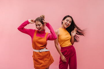 Energetic young two interracial girls dancing side by side and waving hands in studio. Models with light brown and black hair in light spring orange and pink clothes move to music with smile on face.