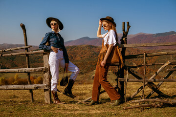 Outdoor full-length fashion portrait of two confident women wearing stylish outfits, posing in autumn mountains. Copy, empty space for text