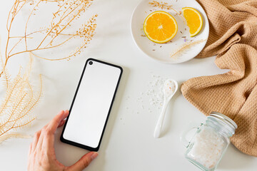 Beauty spa beige composition with 
wooden box, bowl with water, orange slices, blank screen mobile phone and bath salt on white background. Flat lay, top view. Female beauty treatment routine concept.