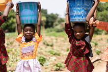 Closeup Of Two Small Black African Girls Carrying Blue Water Buckets On Their Heads; Child Labour Concept
