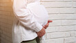© lenblr - Blonde haired pregnant woman in white shirt smiles looking into distance and strokes large tummy standing near white brick wall on sunny day
