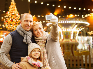 Poster - winter holidays, technology and people concept - happy family taking selfie with camera over evening christmas market or amusement park background