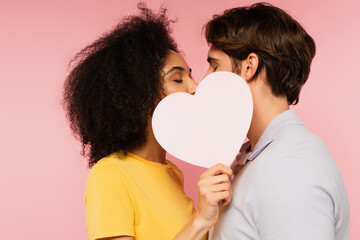 Wall Mural - interracial couple with closed eyes kissing behind blank paper heart isolated on pink.