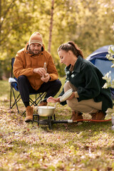 Canvas Print - camping, tourism and travel concept - happy couple drinking beer and cooking food in pot on tourist gas burner at tent camp