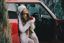 Beautiful Young Woman Wearing Knitted Sweater And Woolen Hat Drinking Hot Chocolate Sitting In Red Car With Christmas Tree On The Top Under Snowfall.