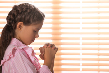 Wall Mural - Cute little girl with hands clasped together praying near window. Space for text